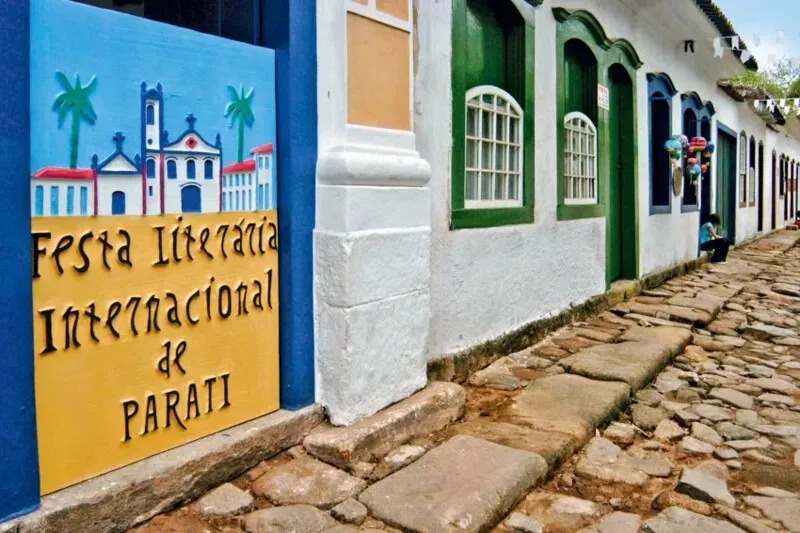 Rua de Paraty em perspectiva com casas coloniais de arede branca e molduras das portas coloridas junto de um painel pintado com a arteda FLIP e rua de pedras redondas